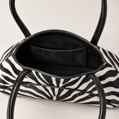 A close-up of a zebra-print handbag with black leather handles and a black interior pocket, shown against a light neutral background.