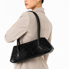 A woman in a beige blazer carries a black crocodile-embossed leather shoulder bag against her back, shown from a side-back angle against a plain white background.