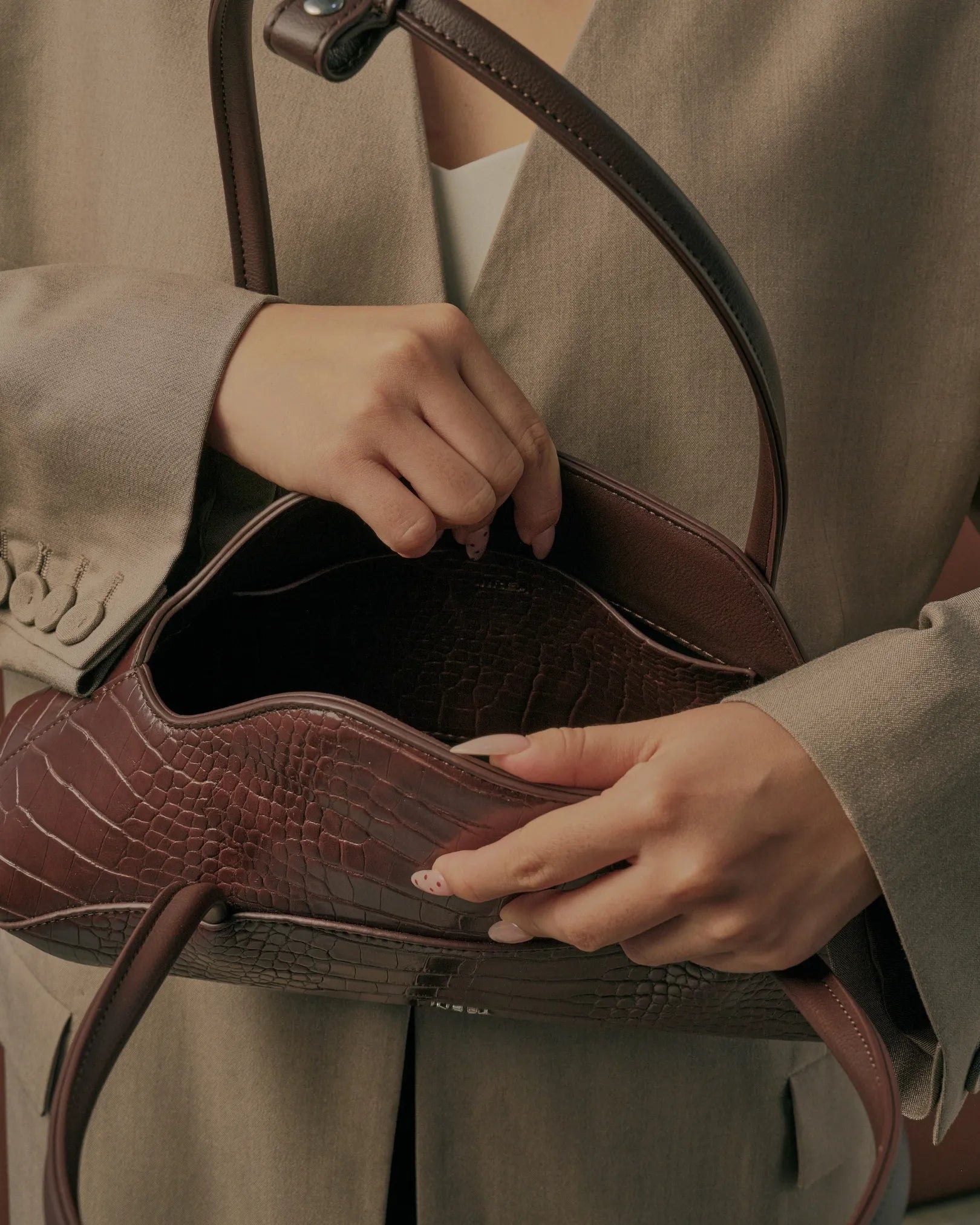 A close-up of a person holding an open brown crocodile-embossed leather handbag, showing the interior lining. The person is wearing a beige blazer.