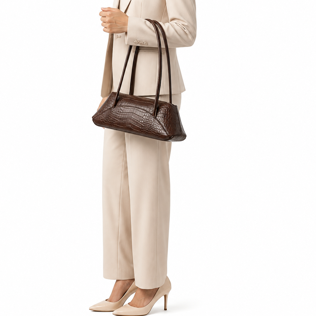 A woman in a beige pantsuit holds a dark brown crocodile-embossed leather shoulder bag against a plain white background.