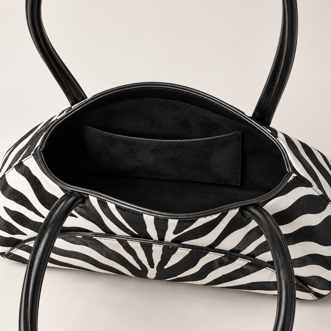 A close-up of a zebra-print handbag with black leather handles and a black interior pocket, shown against a light neutral background.